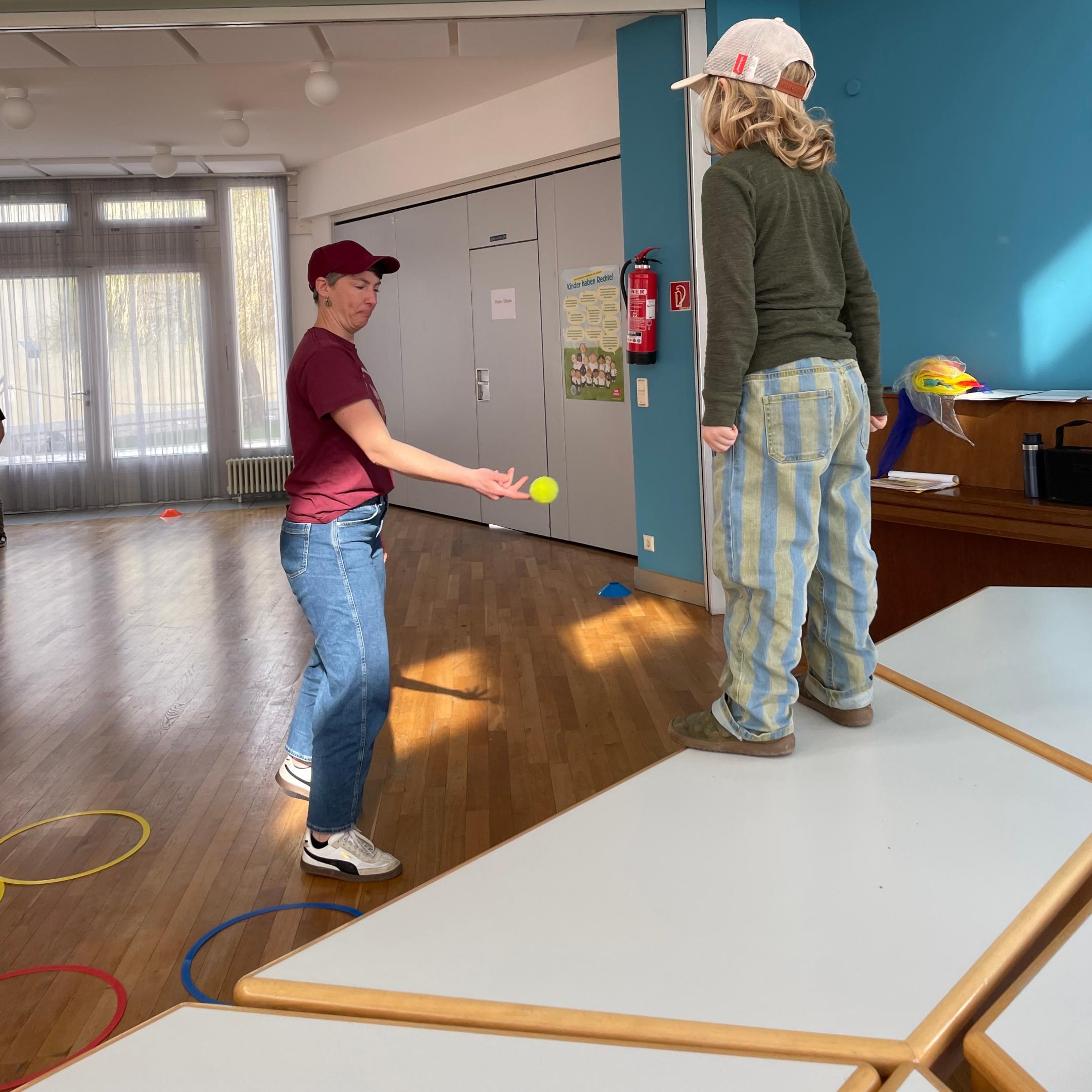 Man sieht ein Kind auf einem Tisch stehend, gegenüber eine Frau die dem Kind einen Tennisball zuwirft