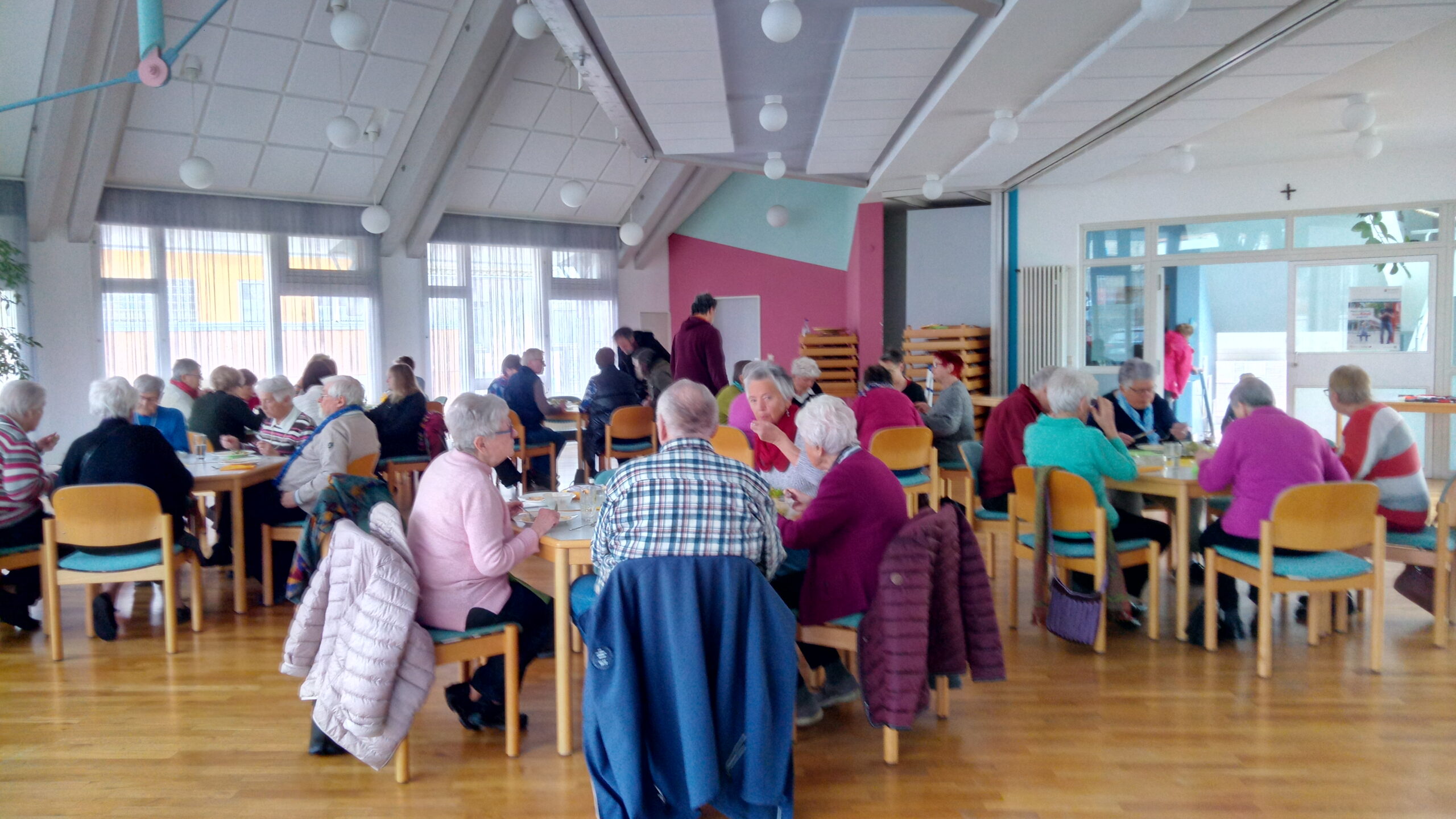 Man sieht die Aula der FaBi mit Personen an mehreren Tischen und Stühle, die dort Mittag essen.