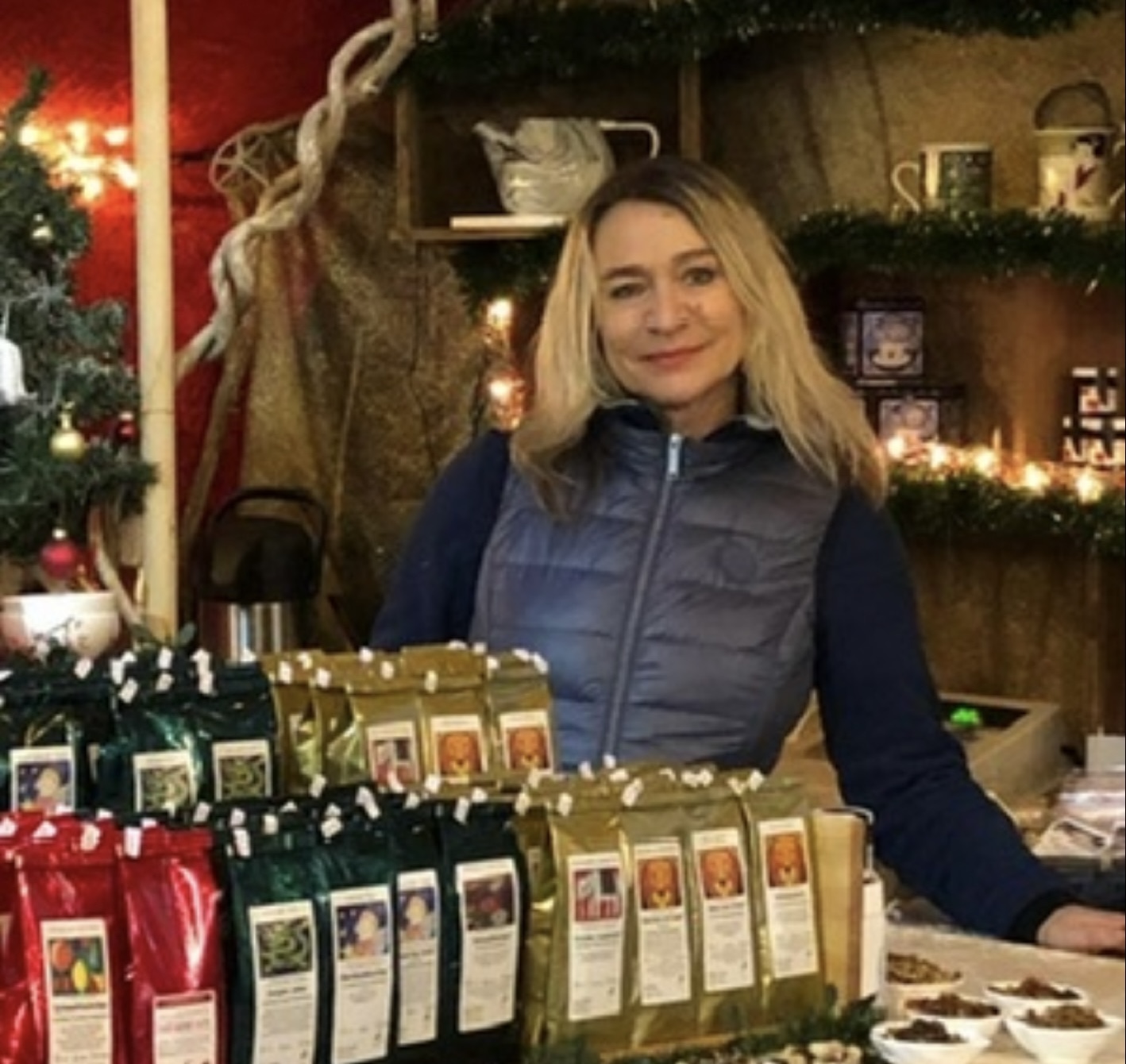 Man sieht eine Frau hinter verschiedene Teesorten. Im Hintergrund sieht man einen geschmückten Tannenbaum, Teekanne und weihnachtliche Lichterkette