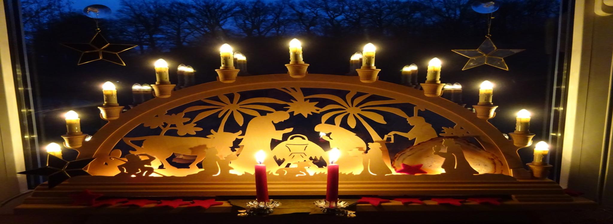 Beleuchteter Schwibbogen mit Weihnachtskrippe und davor brennen 2 rote Kerzen